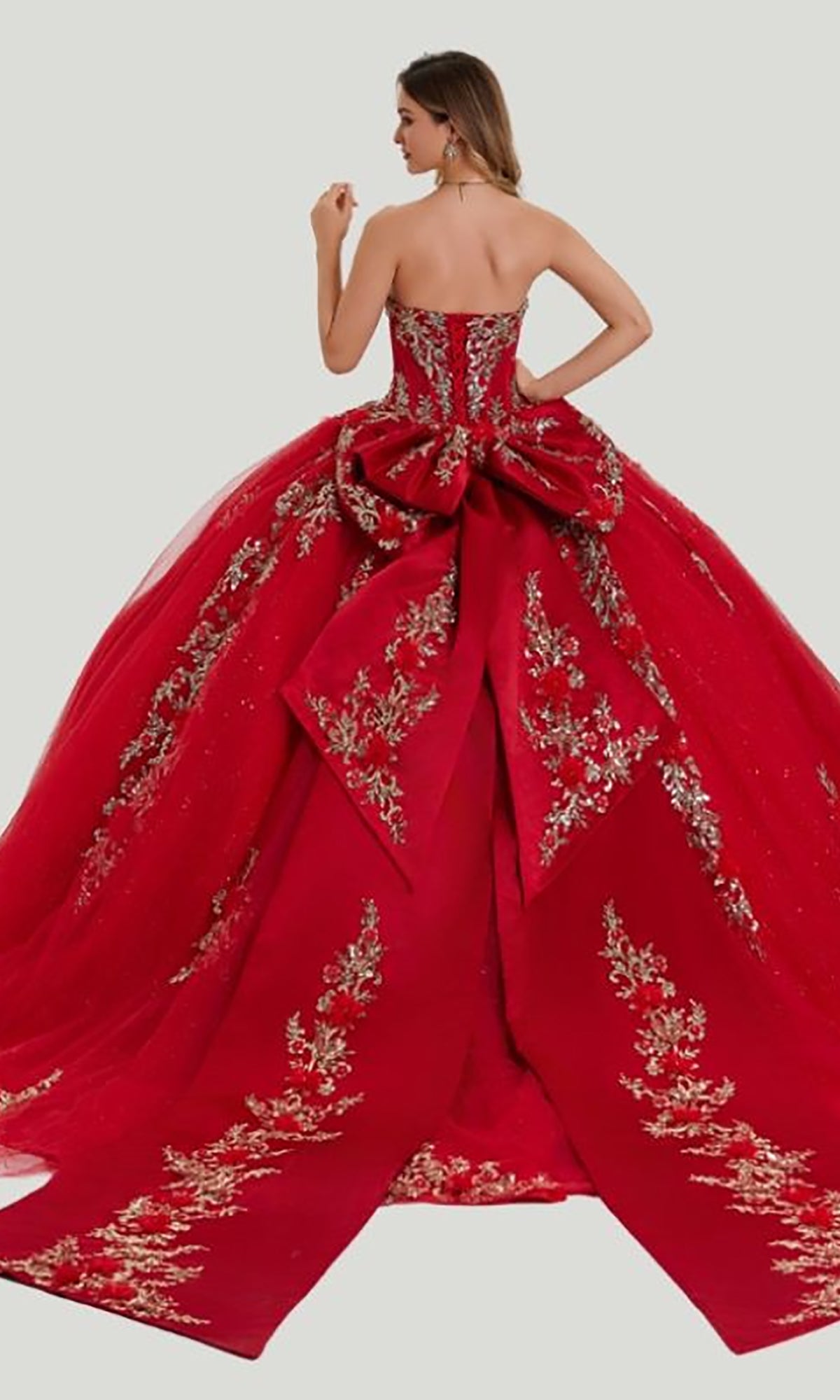 A woman wearing the Quinceanera Dress 26414 by Stella Couture—a strapless red ball gown adorned with gold floral embroidery and embellished appliques, featuring a large bow at the back—stands against a plain light background.